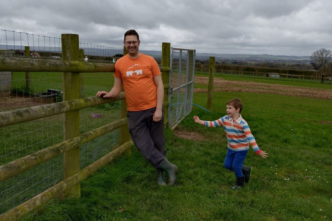 Tony from SeaNic wearing a hand-printed orange organic cotton t-shirt, leaning on a wooden fence while his son Arthur plays in a green field with the Haldon Hills in Devon in the background.