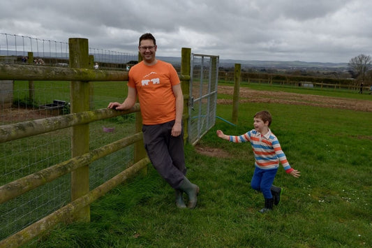 Tony from SeaNic wearing a hand-printed orange organic cotton t-shirt, leaning on a wooden fence while his son Arthur plays in a green field with the Haldon Hills in Devon in the background.