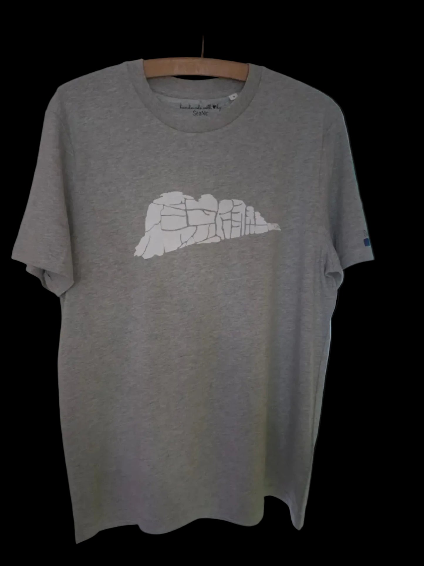 A grey organic cotton T-shirt by SeaNic, featuring a white screen-printed graphic of Haytor Rocks on Dartmoor. The shirt is displayed on a vintage wooden hanger against a neutral background.
