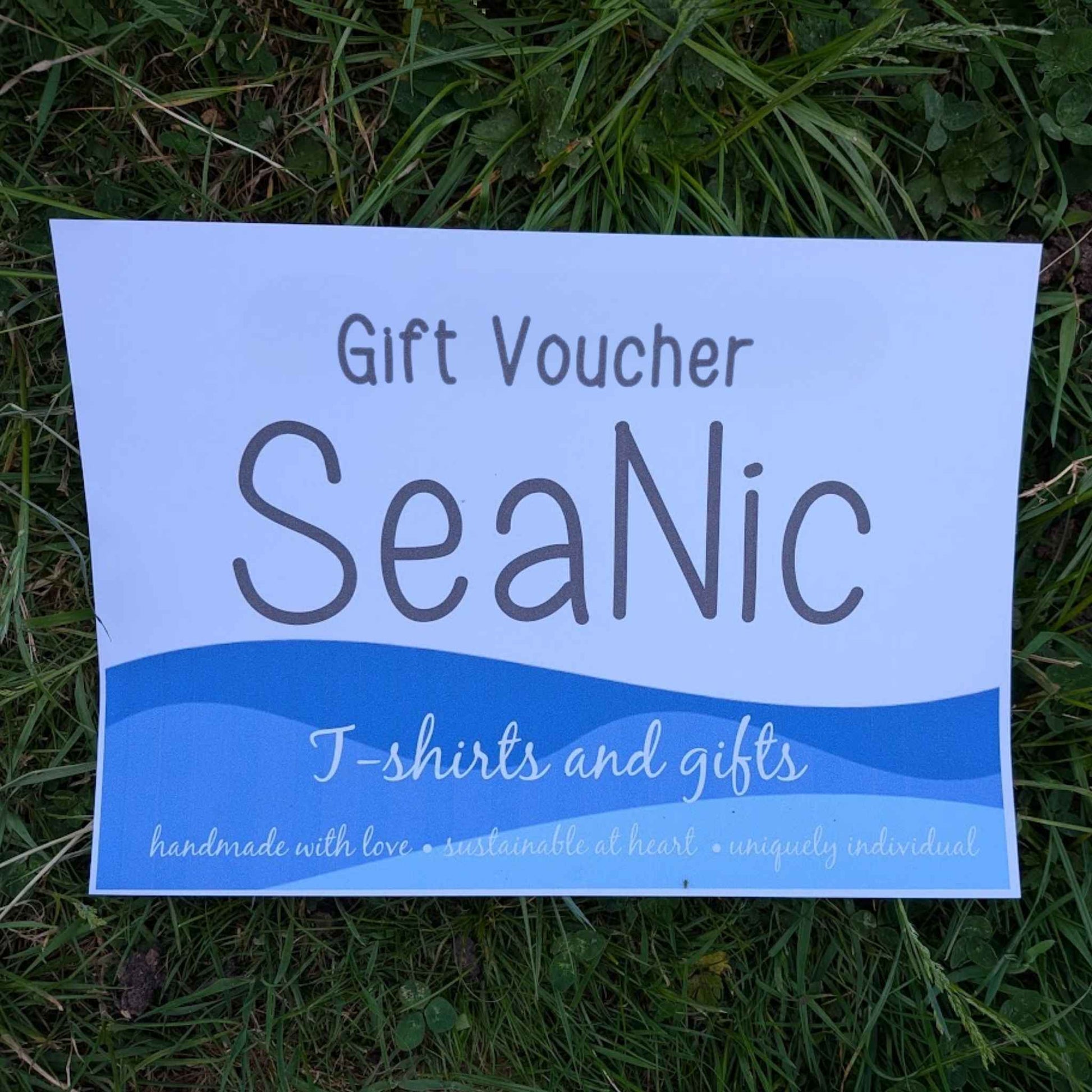 SeaNic gift voucher card laid on grass, featuring the SeaNic logo, wave design in blue and white, and the straplines handmade with love, sustainable at heart, uniquely individual.